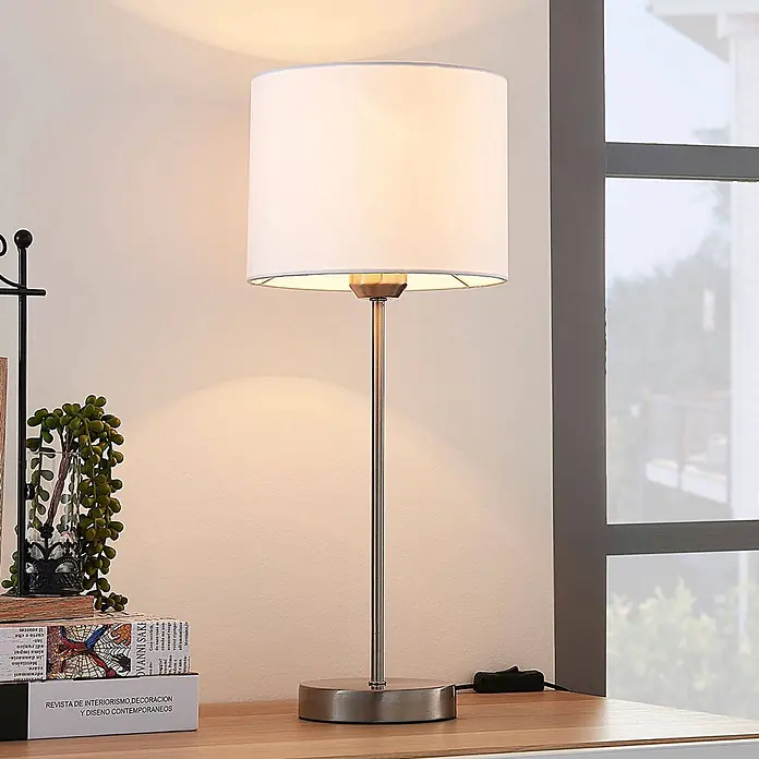Lindby Tischleuchte Annatina - L x B x H: 20 x 20 x 48 cm, alu / grau / zink, weiß / opal, E27 - Elegante Tischlampe mit weißem Stoffschirm und gebürstetem silberfarbenem Metallsockel und -stiel, die ein warmes Licht auf einen Holzschreibtisch wirft.