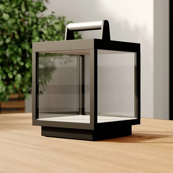 Lucande Tischleuchte Cube - schwarz - Moderne anthrazitfarbene Außenlaterne mit klaren Glaspaneelen, silbernem Griff und hellem Boden auf einem Holzdeck.