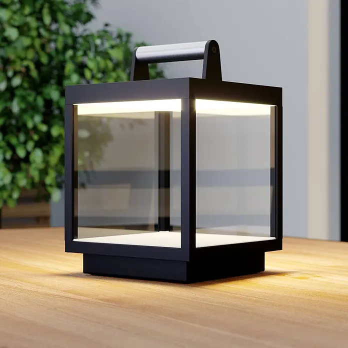 Lucande Tischleuchte Cube - schwarz - Schwarze moderne Laterne mit Klarglas und silbernem Griff, leuchtend auf einem Holztisch.
