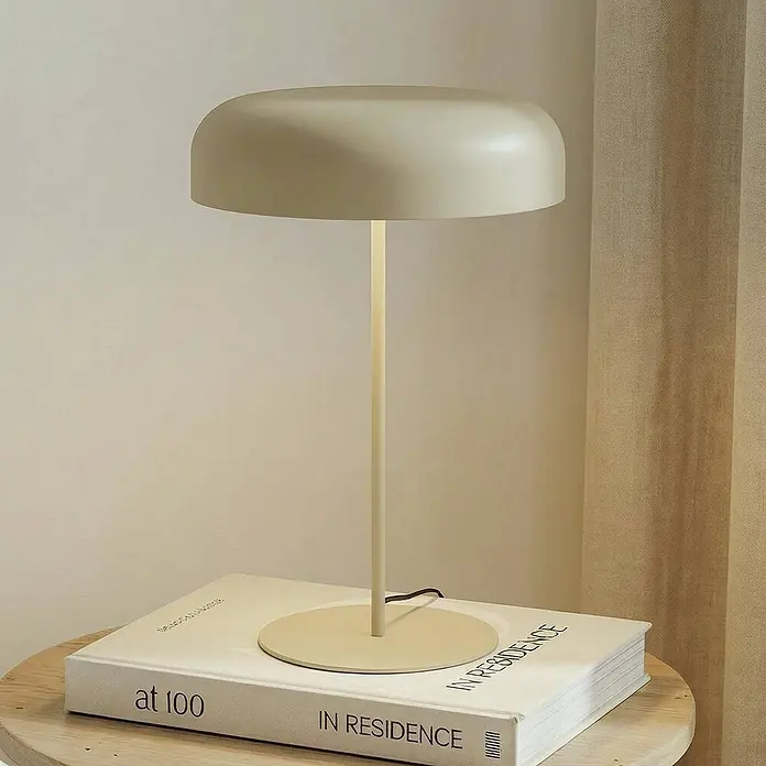 Lindby Tischleuchte Edion - braun / rost, braun / rost, E14 - Eine cremefarbene, minimalistische Tischleuchte mit pilzförmigem Schirm steht auf einem Buch auf einem Holztisch.