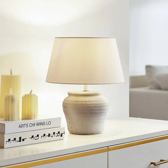 Lindby Tischleuchte Aelith - L x B x H: 30 x 30 x 36 cm, weiß / opal, weiß / opal, E27 - Strukturierte cremeweiße Keramiktischlampe mit hellem Stoffschirm auf einem weißen Schrank, neben Kerzen und Büchern.