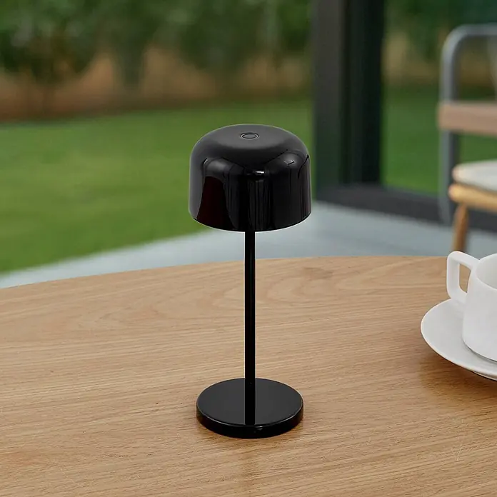 Lucande Tischleuchte Quill - schwarz, schwarz - Glänzende schwarze tragbare Lampe auf einem hellen Holztisch im Freien.