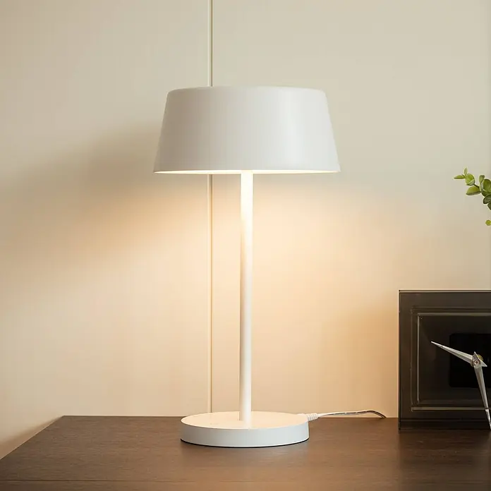 Lindby Tischleuchte Milica - L x B x H: 18,3 x 18,3 x 33,6 cm, weiß / opal, weiß / opal - Beleuchtete weiße Tischlampe mit zylindrischem Schirm, auf einer dunklen Holzoberfläche stehend.