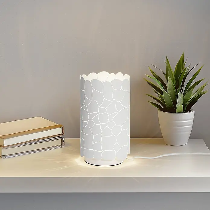 Lindby Tischleuchte Aralena - weiß / opal, E27 - Weiße Tischlampe mit dekorativem gemustertem Schirm, wirft Licht auf ein Regal neben Büchern und einer Pflanze.