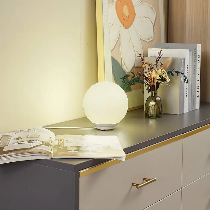 Lindby Tischleuchte Cosmia - L x B x H: 15 x 15 x 15,7 cm, weiß / opal - Eine beleuchtete kugelförmige Tischlampe mit gefrosteter weißer Kugel und weißem Sockel auf einer Kommode.