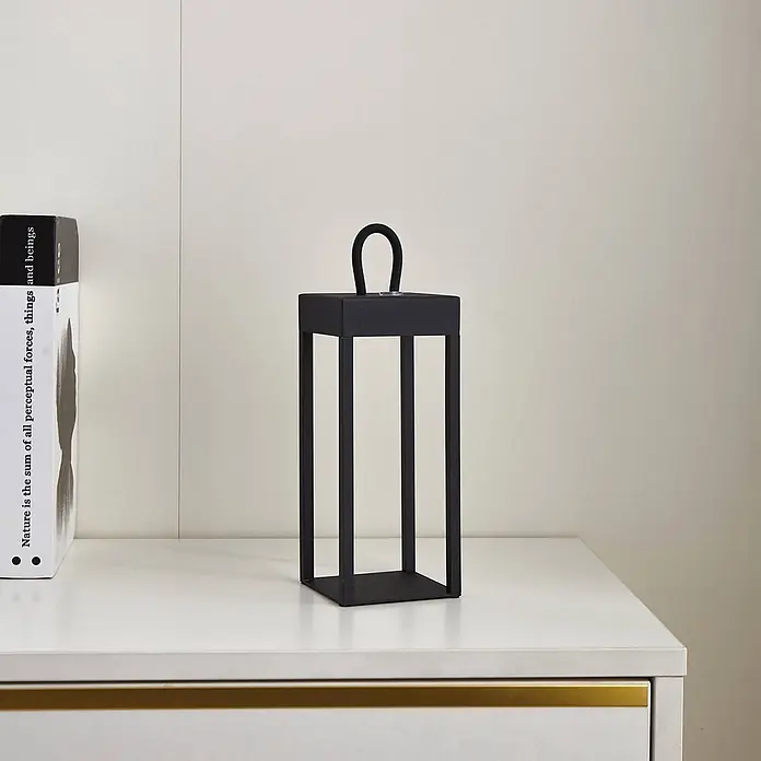 Lindby Tischleuchte Iruna - L x B x H: 9 x 9 x 26,5 cm, schwarz, schwarz - Schwarze minimalistische Laterne mit Griff auf einer weißen Oberfläche neben einem Buch.