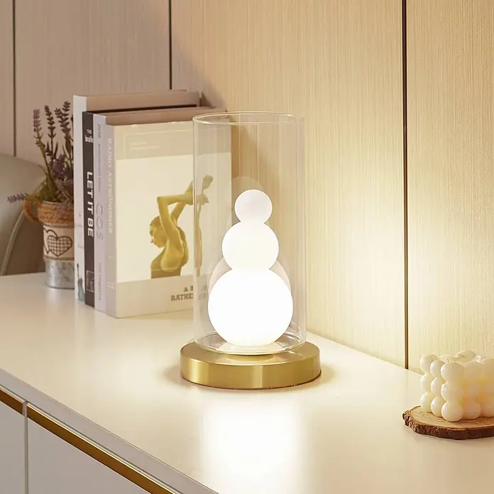 Lindby Tischleuchte Kupla - L x B x H: 15 x 15 x 28,5 cm, messing / gold, klar / transparent, G9 - Moderne Tischleuchte mit Messingsockel, klarem Glaszylinder und drei leuchtenden weißen Kugeln, beleuchtet eine helle Oberfläche.