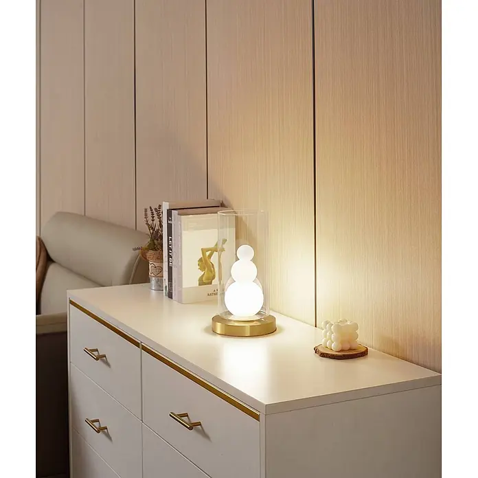 Lindby Tischleuchte Kupla - L x B x H: 15 x 15 x 28,5 cm, messing / gold, klar / transparent, G9 - Elegante Tischleuchte mit klarem Glaszylinder, drei weißen Kugelleuchten auf goldenem Sockel, auf einer Kommode.