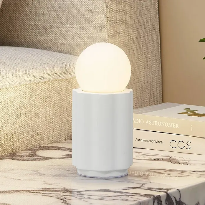 Lindby Tischleuchte Nielson - L x B x H: 11 x 11 x 23 cm, weiß / opal, weiß / opal, E27 - Eine beleuchtete weiße kugelförmige Tischlampe mit gerilltem zylindrischem Sockel steht auf einer Marmoroberfläche neben Büchern und einem Sofa.