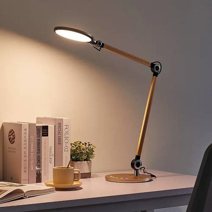 Lindby Tischleuchte Nyxaris - L x B x H: 55,7 x 20,1 x 49,7 cm, messing / gold, messing / gold - Moderne Schreibtischlampe mit einem gelenkigen Holzarm und einem hellen LED-Kopf, die einen Schreibtisch mit Büchern und einer Tasse beleuchtet.