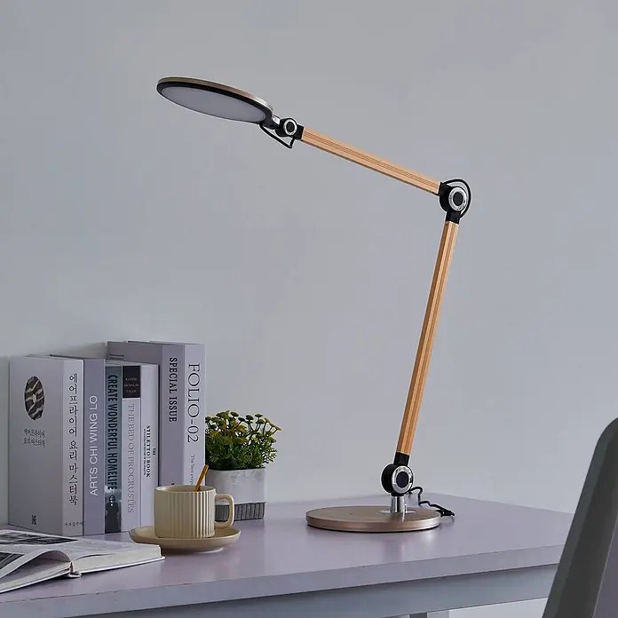 Lindby Tischleuchte Nyxaris - L x B x H: 55,7 x 20,1 x 49,7 cm, messing / gold, messing / gold - Moderne Schreibtischlampe mit hellem Holzgelenkarm, schwarzen Gelenken und goldumrandetem LED-Kopf, auf einem Schreibtisch mit Büchern und Pflanze.