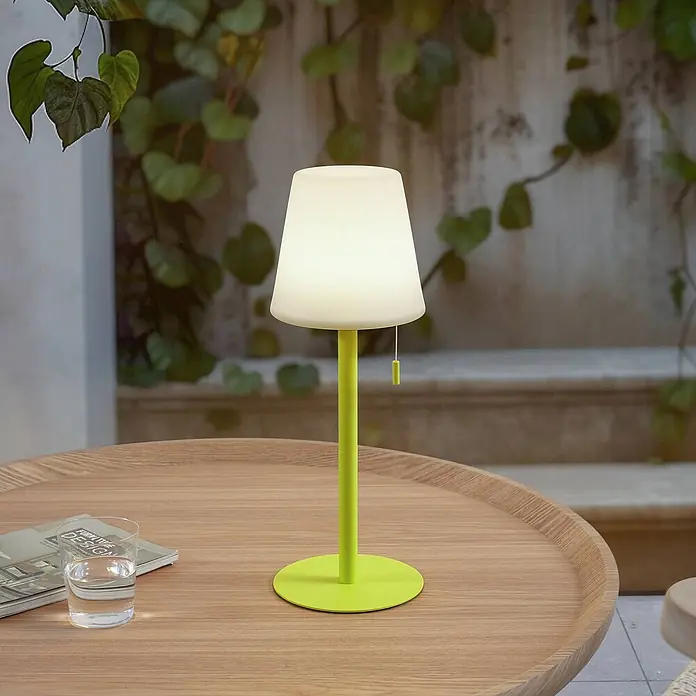 Lindby Tischleuchte Azalea - gelb / orange - Limettengrüne Tischlampe mit weißem Schirm auf einem Holztisch im Freien.