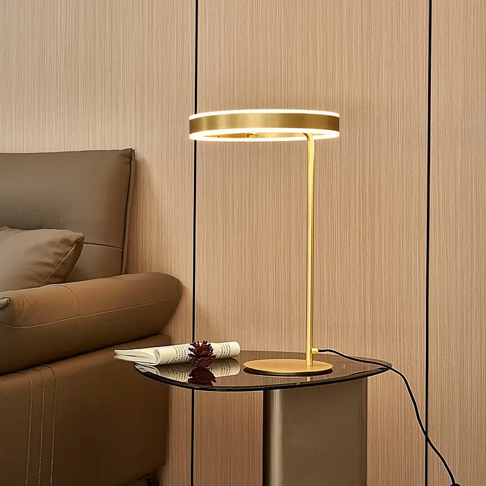 Lucande Tischleuchte Yekta - L x B x H: 30 x 30 x 58,5 cm, messing / gold, messing / gold - Moderne goldfarbene Tischlampe mit beleuchtetem Rundkopf, auf einem Glastisch neben einem braunen Sessel.