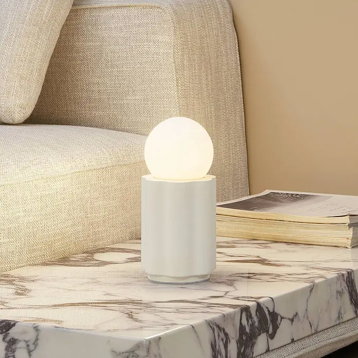 Lindby Tischleuchte Nielson - creme / amber, weiß / opal, E27 - Eine weiße zylindrische Tischlampe mit kugelförmigem Licht auf einem marmorierten Beistelltisch neben einem beigen Sofa.