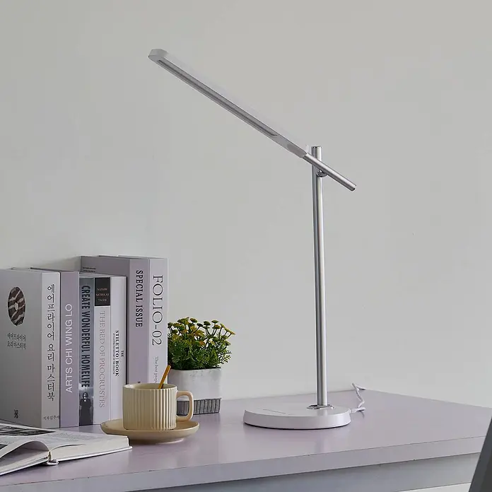 Lindby Tischleuchte Valtorin - weiß / opal, weiß / opal - Eine moderne silber-weiße Schreibtischlampe mit verstellbarem Leuchtbalken auf einem Schreibtisch neben Büchern und einer Pflanze.