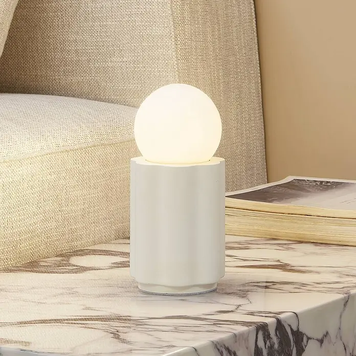Lindby Tischleuchte Nielson - creme / amber, weiß / opal, E27 - Weiße Keramik-Tischleuchte mit kugelförmigem Diffusor, sanftes Licht spendend, auf einem Marmor-Beistelltisch neben einem beigen Sofa.