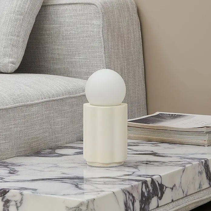 Lindby Tischleuchte Nielson - creme / amber, weiß / opal, E27 - Cremefarbene Tischleuchte mit kugelförmigem weißem Diffusor auf Marmor-Beistelltisch neben grauem Sofa.