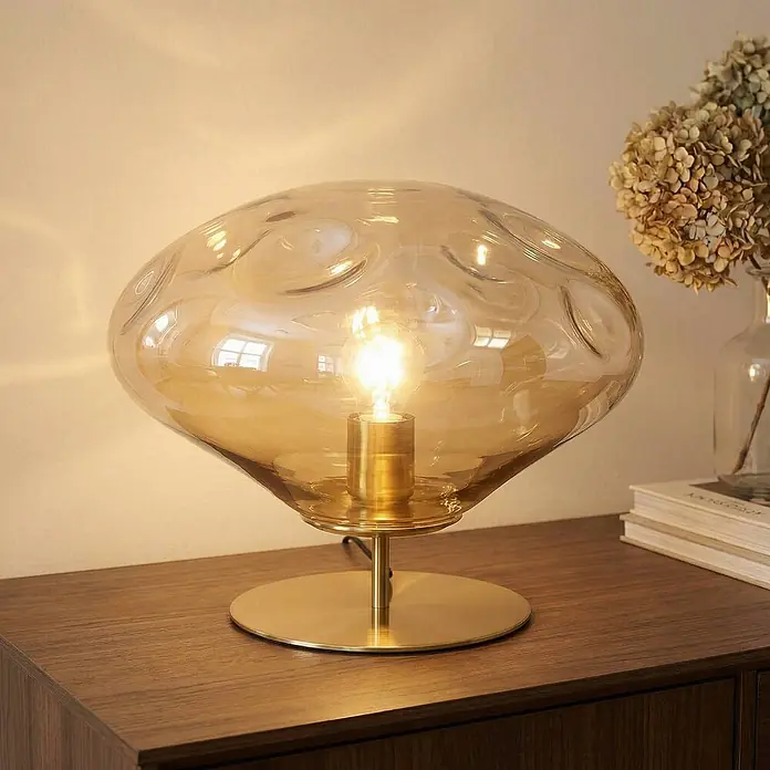 Lindby Tischleuchte Lelia - L x B x H: 35 x 35 x 27,5 cm, creme / amber, E27 - Beleuchtete Tischleuchte mit bernsteinfarbenem Wellenglas und Messingsockel auf Holzfläche.