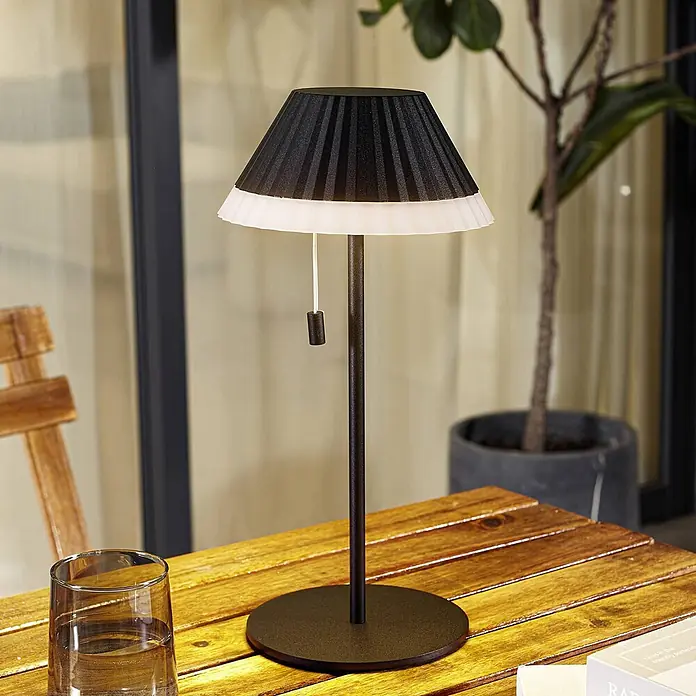 Lindby Tischleuchte Ilvi - L x B x H: 17,5 x 17,5 x 35 cm, schwarz, schwarz - Eine moderne schwarz-weiße Falten-Tischleuchte mit dunklem Metallschaft steht auf einem Lattenholztisch im Freien.