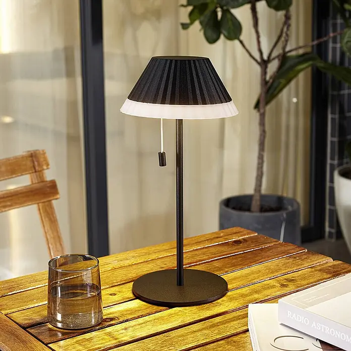 Lindby Tischleuchte Ilvi - L x B x H: 17,5 x 17,5 x 35 cm, schwarz, schwarz - Schwarze plissierte Tischlampe mit weißem Faltenrand auf dunklem Fuß, auf einem Holztisch.