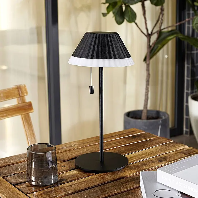 Lindby Tischleuchte Ilvi - L x B x H: 17,5 x 17,5 x 35 cm, schwarz, schwarz - Schwarze Tischlampe mit plissiertem Schirm und weißem Rand, auf einem Holztisch im Freien.