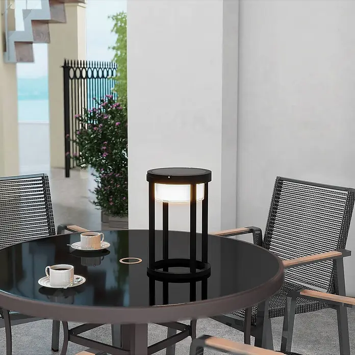 Lucande Tischleuchte Tilena - L x B x H: 21 x 21 x 32 cm, schwarz - Schwarze Outdoor-Tischleuchte mit beleuchtetem Diffusor, auf einem runden Glastisch mit Kaffeetassen platziert.