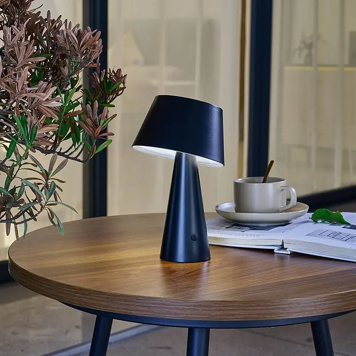 Lindby Tischleuchte Lirinor - L x B x H: 13,16 x 13,16 x 21,8 cm, schwarz, schwarz - Schwarze tragbare Tischlampe mit konischem Fuß und schrägem Schirm, beleuchtet, auf Holztisch mit Pflanze, Buch und Tasse.