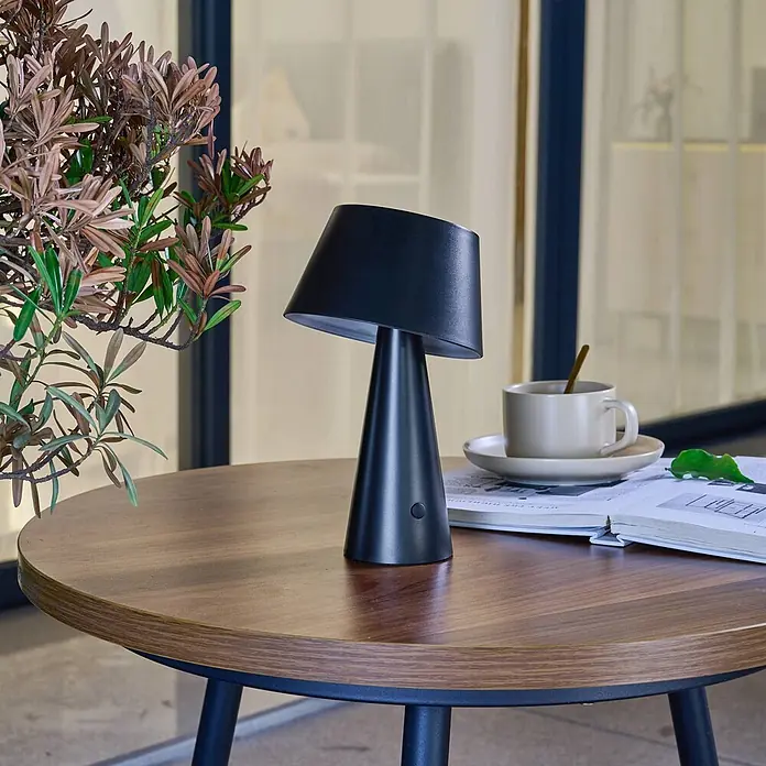 Lindby Tischleuchte Lirinor - L x B x H: 13,16 x 13,16 x 21,8 cm, schwarz, schwarz - Schwarze tragbare Tischlampe auf einem Holztisch, neben einer Kaffeetasse und einem offenen Buch.