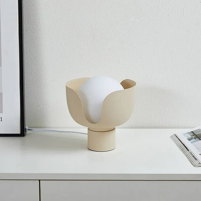 Lindby Tischleuchte Fiora - L x B x H: 20 x 20 x 21 cm, creme / amber, E27 - Eine beigefarbene tulpenförmige Tischleuchte mit kugelförmigem weißen Diffusor steht neben einem Bilderrahmen und Zeitschriften.