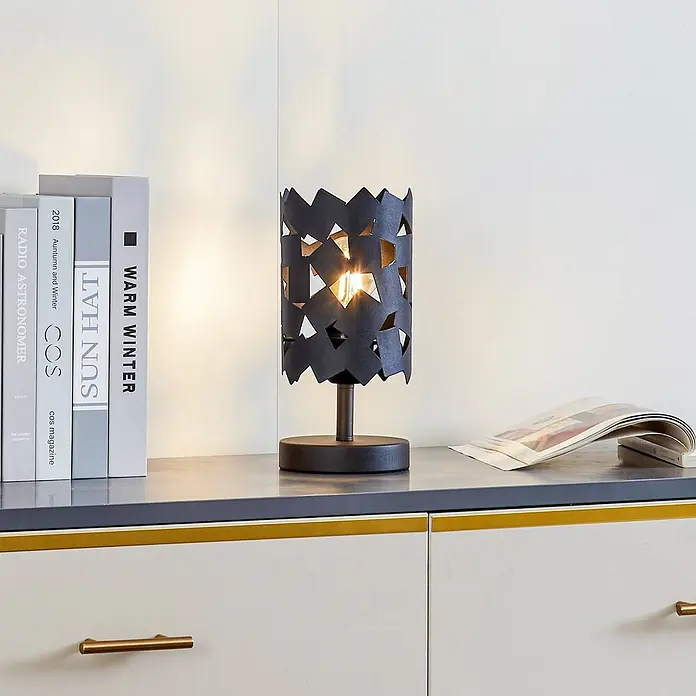 Lucande Tischleuchte Aeloria - L x B x H: 12 x 12 x 25 cm, schwarz, schwarz, E27 - Dunkle Metall-Tischlampe mit geometrisch ausgeschnittenem Schirm, leuchtend auf einem Schrank neben Büchern.