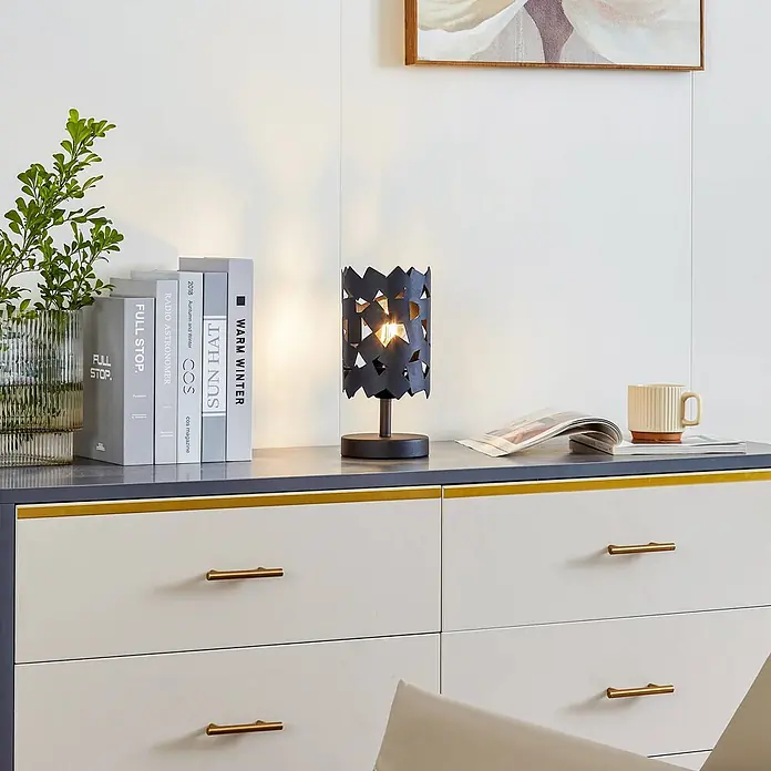 Lucande Tischleuchte Aeloria - L x B x H: 12 x 12 x 25 cm, schwarz, schwarz, E27 - Eine schwarze Tischlampe mit ausgeschnittenem geometrischem Metallschirm steht auf einer Kommode und wirft komplexe Lichtmuster.