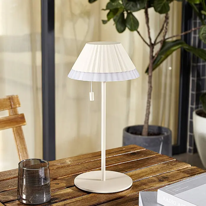 Lindby Tischleuchte Ilvi - L x B x H: 17,5 x 17,5 x 35 cm, creme / amber, creme / amber - Cremefarbene Tischleuchte mit plissiertem Schirm und weißer Rüsche, auf einem Holztisch im Freien gezeigt.