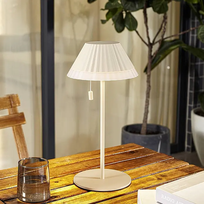 Lindby Tischleuchte Ilvi - L x B x H: 17,5 x 17,5 x 35 cm, creme / amber, creme / amber - Eine elegante cremefarbene Tischleuchte mit plissiertem Stoffschirm steht auf einem Holztisch im Freien neben einem Glas Wasser.