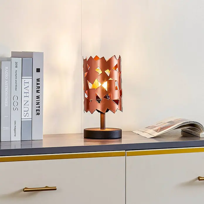 Lucande Tischleuchte Aeloria - L x B x H: 12 x 12 x 25 cm, kupfer, E27 - Kupferfarbene Metall-Tischleuchte mit geometrischen Aussparungen, beleuchtet ein Regal neben Büchern.
