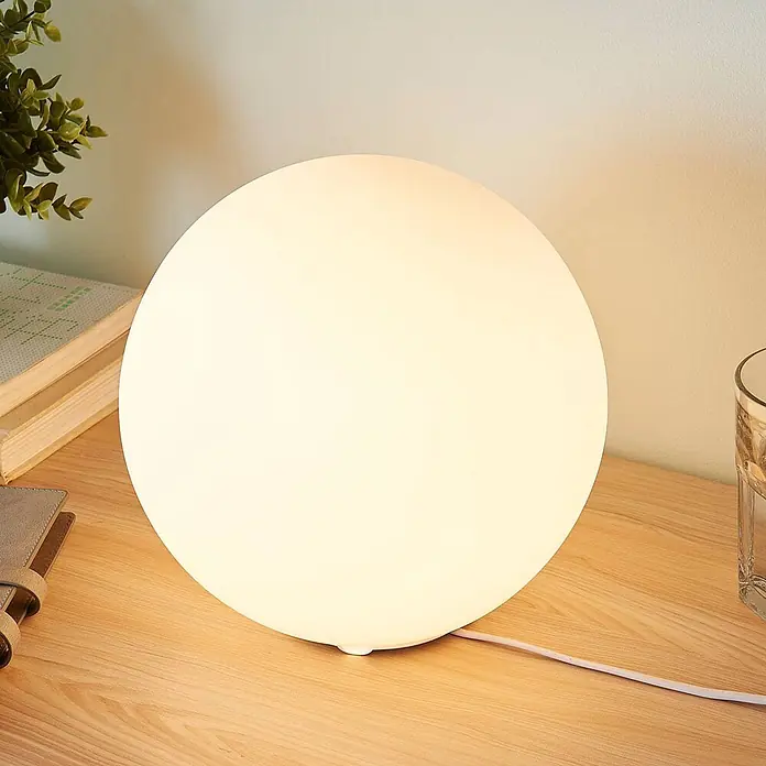 Lindby Tischleuchte Marike - L x B x H: 25 x 25 x 24,5 cm, weiß / opal, E27 - Eine leuchtende, milchweiße kugelförmige Tischlampe steht auf einem Holzschreibtisch und spendet warmes Licht.