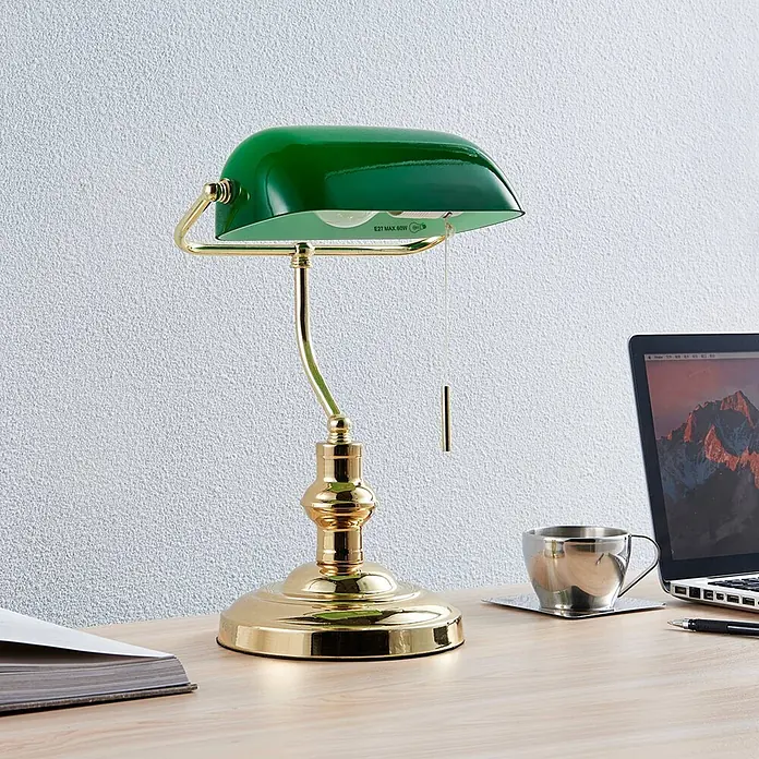 Lindby Tischleuchte Milenka - L x B x H: 22,5 x 25,8 x 39 cm, messing / gold, grün, E27 - Grüne und goldfarbene Bankerlampe auf einem Holzschreibtisch mit Laptop und Kaffeetasse.