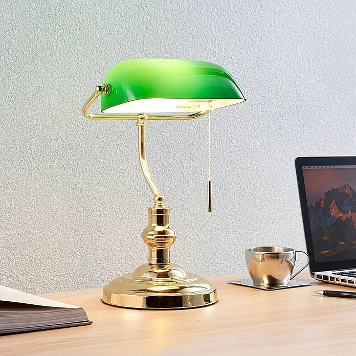 Lindby Tischleuchte Milenka - L x B x H: 22,5 x 25,8 x 39 cm, messing / gold, grün, E27 - Eine beleuchtete Messing-Bankerlampe mit grünem Glasschirm auf einem hellen Holzschreibtisch neben Laptop und Tasse.