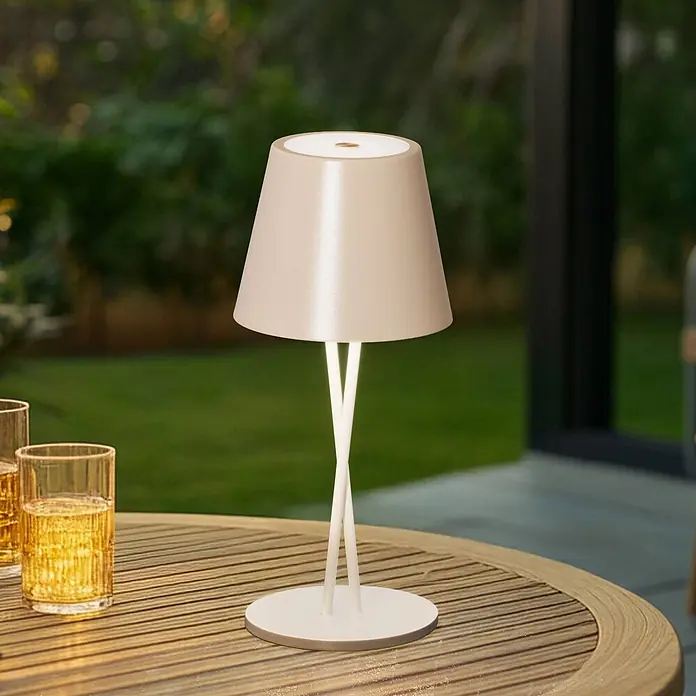 Lindby Tischleuchte Janea - creme / amber, creme / amber - Eine kabellose beigefarbene Tischleuchte mit konischem Schirm und überkreuzten Beinen steht auf einem Holztisch im Freien neben zwei Gläsern.