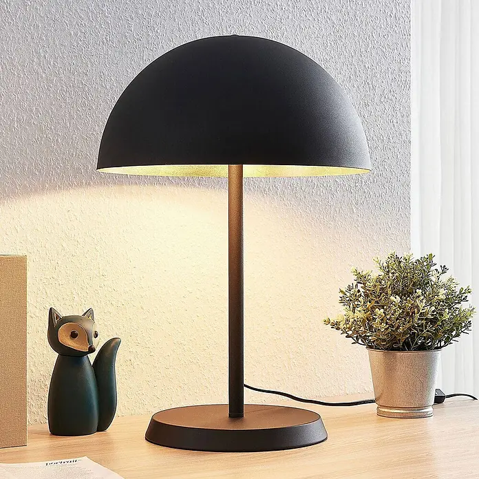 Lindby Tischleuchte Idalene - L x B x H: 35 x 35 x 51 cm, schwarz, schwarz, E27 - Schwarze Kuppel-Tischleuchte mit goldenem Innenraum, leuchtend auf einem Holzschreibtisch.