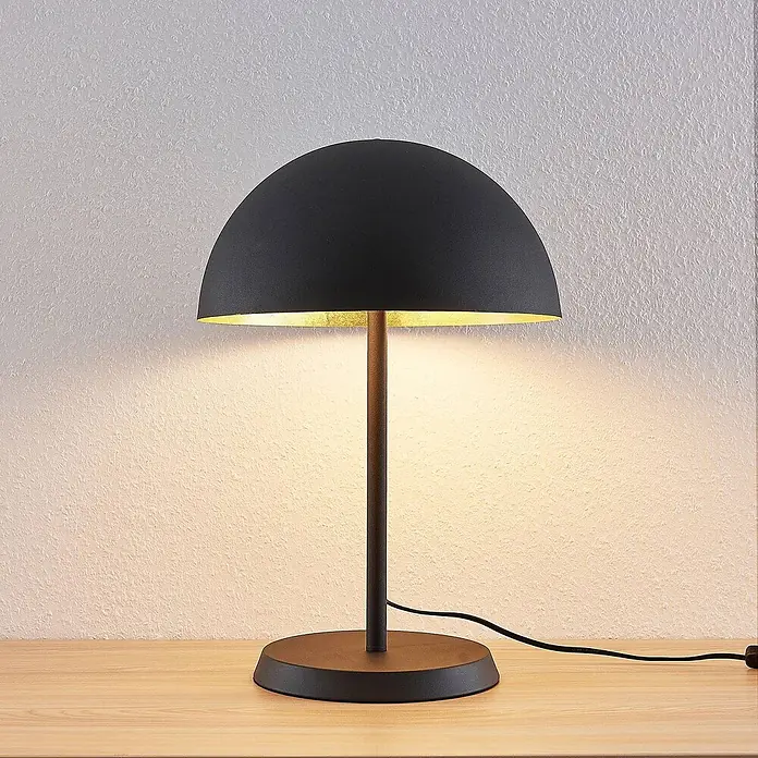 Lindby Tischleuchte Idalene - L x B x H: 35 x 35 x 51 cm, schwarz, schwarz, E27 - Schwarze Tischlampe mit kuppelförmigem Schirm und goldener Innenseite, spendet warmes Licht auf eine Wand, auf Holztisch.