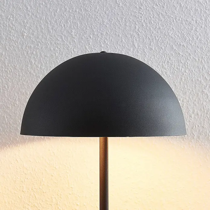 Lindby Tischleuchte Idalene - L x B x H: 35 x 35 x 51 cm, schwarz, schwarz, E27 - Mattschwarzer halbkuppelförmiger Lampenschirm auf dunklem Metallfuß, der warmes Licht auf eine strukturierte Wand wirft.
