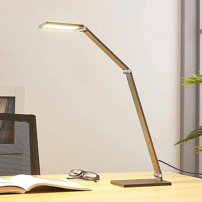 Lucande Tischleuchte Mion - L x B x H: 36,5 x 12 x 41 cm, alu / grau / zink, alu / grau / zink - Moderne bronzefarbene LED-Schreibtischlampe mit verstellbarem Arm auf einem Holzschreibtisch mit einem Buch und Brille.