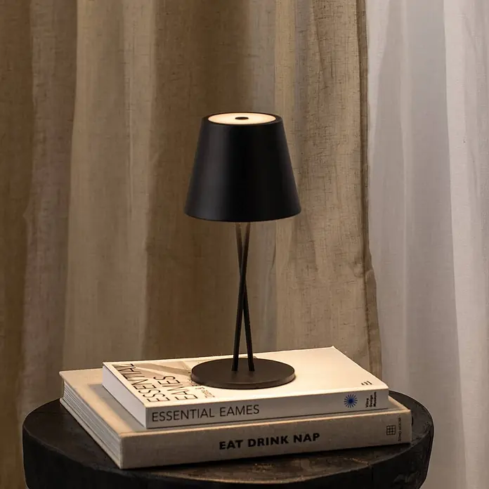 Lindby Tischleuchte Janea - L x B x H: 11,4 x 11,4 x 25 cm, schwarz, schwarz - Eine schwarze Tischlampe mit konischem Schirm und dünnen gekreuzten Beinen steht auf einem Bücherstapel vor hellen Vorhängen.