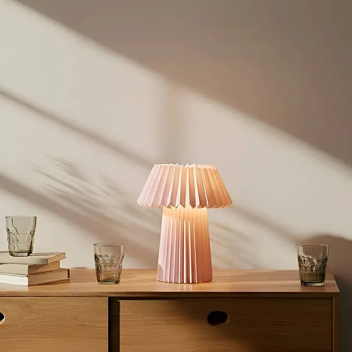 Lindby Tischleuchte Magali - L x B x H: 34 x 34 x 37 cm, violett / pink / rose, E14 - Eine pinkfarbene plissierte Tischlampe beleuchtet eine Holzkommode, Licht strömt aus Schirm und Sockel. Bücher und Gläser stehen daneben.