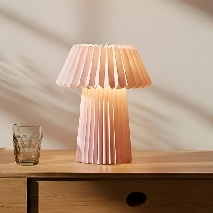 Lindby Tischleuchte Magali - L x B x H: 34 x 34 x 37 cm, violett / pink / rose, E14 - Rosafarbene plissierte Tischlampe auf einem Holztisch, die warmes Licht ausstrahlt.