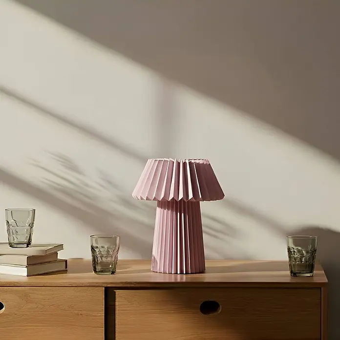 Lindby Tischleuchte Magali - L x B x H: 34 x 34 x 37 cm, violett / pink / rose, E14 - Eine rosa plissierte Tischlampe steht auf einer Holzkommode, umgeben von Gläsern und Büchern.