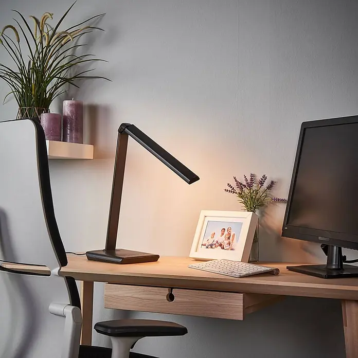 Lindby Tischleuchte Kuno - L x B x H: 37,6 x 15,8 x 46 cm, schwarz, schwarz - Schwarze Schreibtischlampe mit verstellbarem Arm, die einen Holztisch mit Computermonitor, Tastatur und gerahmtem Foto beleuchtet.