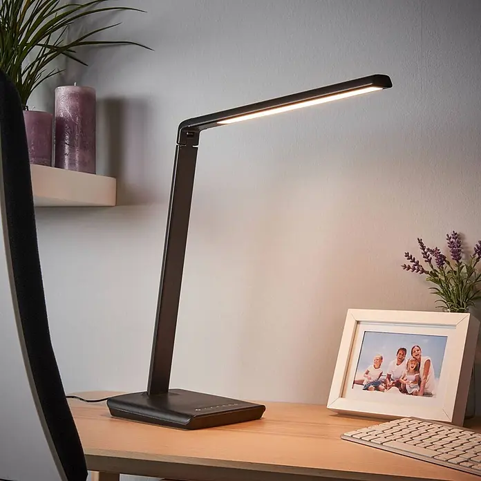 Lindby Tischleuchte Kuno - L x B x H: 37,6 x 15,8 x 46 cm, schwarz, schwarz - Schwarze LED-Schreibtischlampe mit rechteckigem Kopf, leuchtend, auf einem Holzschreibtisch neben einer Tastatur und einem gerahmten Foto.