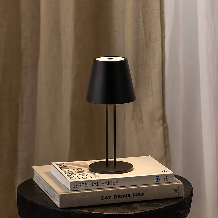 Lindby Tischleuchte Janea - L x B x H: 11,4 x 11,4 x 25 cm, schwarz, schwarz - Schwarze tragbare Tischlampe mit konischem Schirm auf einem Stapel Bücher auf einem dunklen Holzhocker.