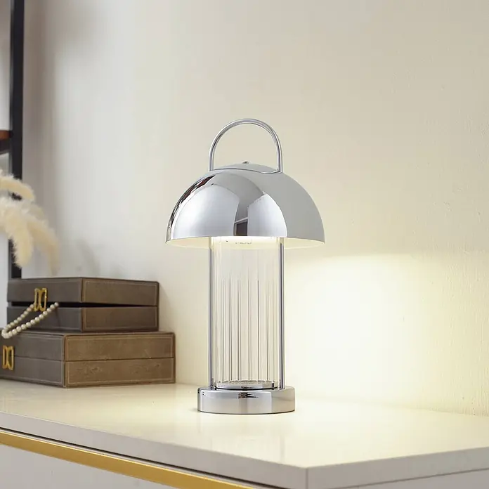 Lindby Tischleuchte Mareli - klar / transparent, chrom / silber - Polierte Chrom-Tischleuchte mit gewölbtem Schirm, Griff und geripptem Glas, beleuchtet auf einer Ablage.